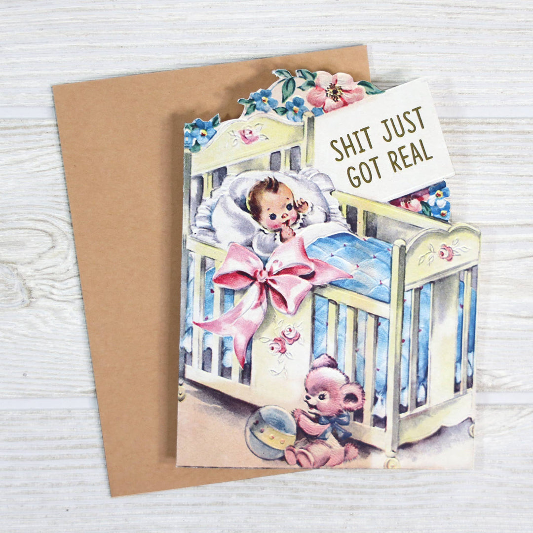 Card with a baby in a crib and a pink bow, featuring a humorous message "SHIT JUST GOT REAL."
