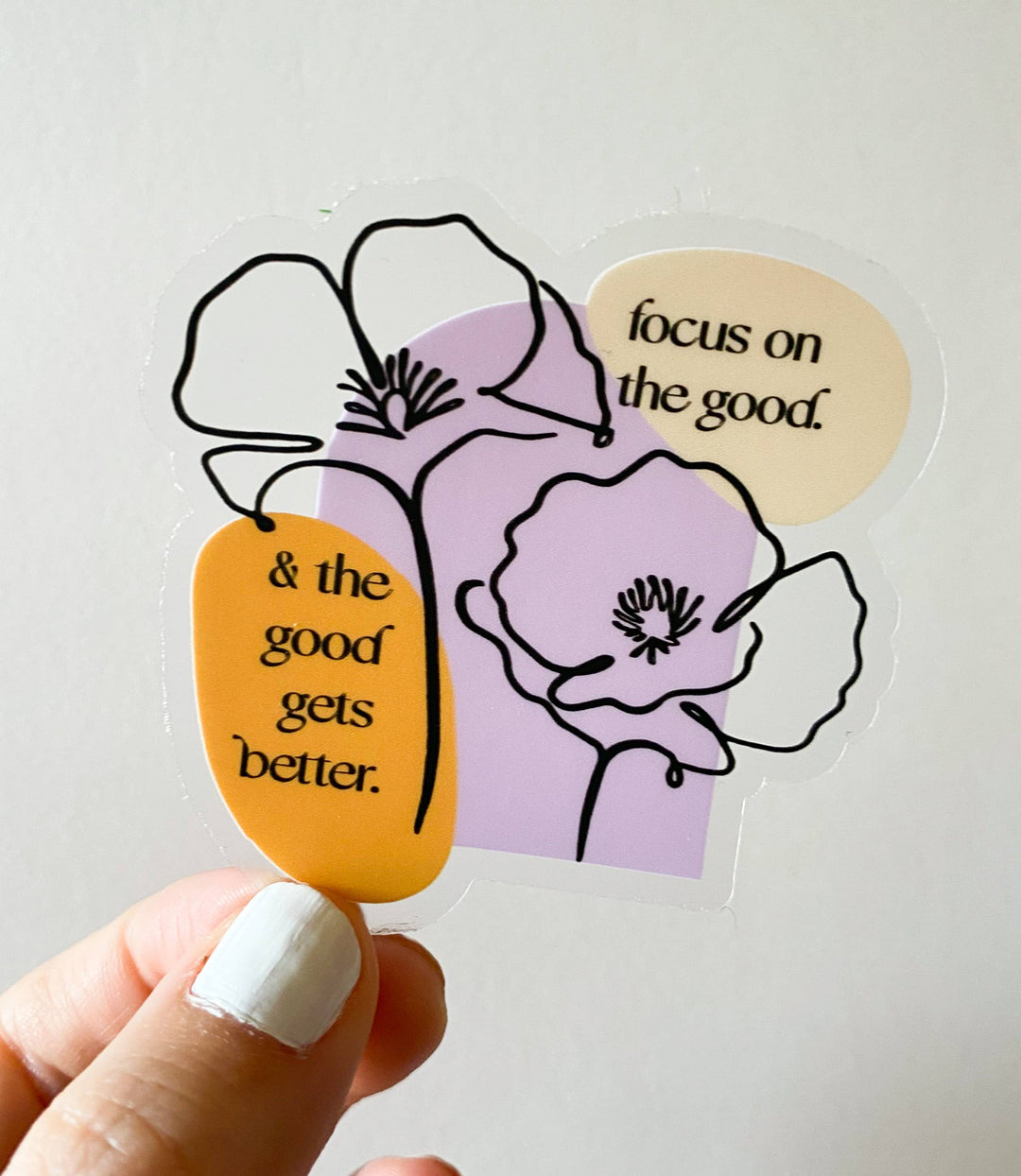 A hand with white nail polish holds a die-cut sticker featuring black line art of flowers and two quote bubbles, one orange and one cream, over a lavender background. 