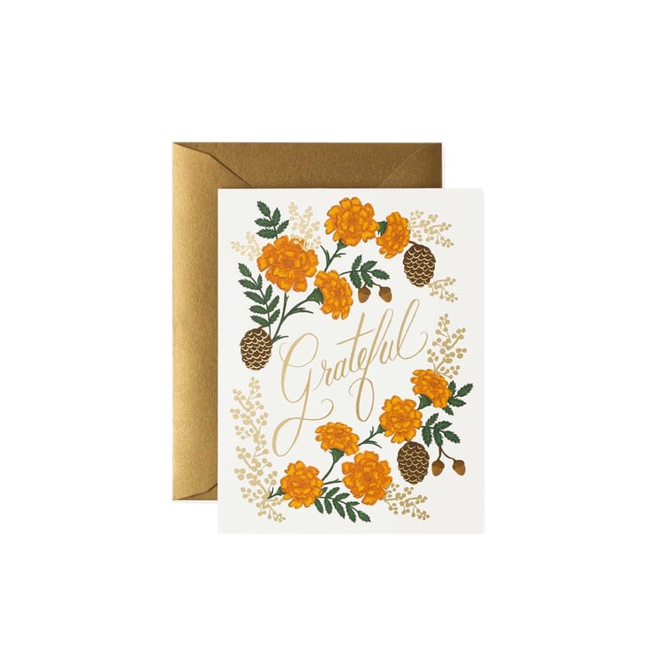 A white greeting card with a gold envelope behind it, featuring an illustration of orange marigolds, green leaves, pinecones, and the word 'Grateful' in gold script.