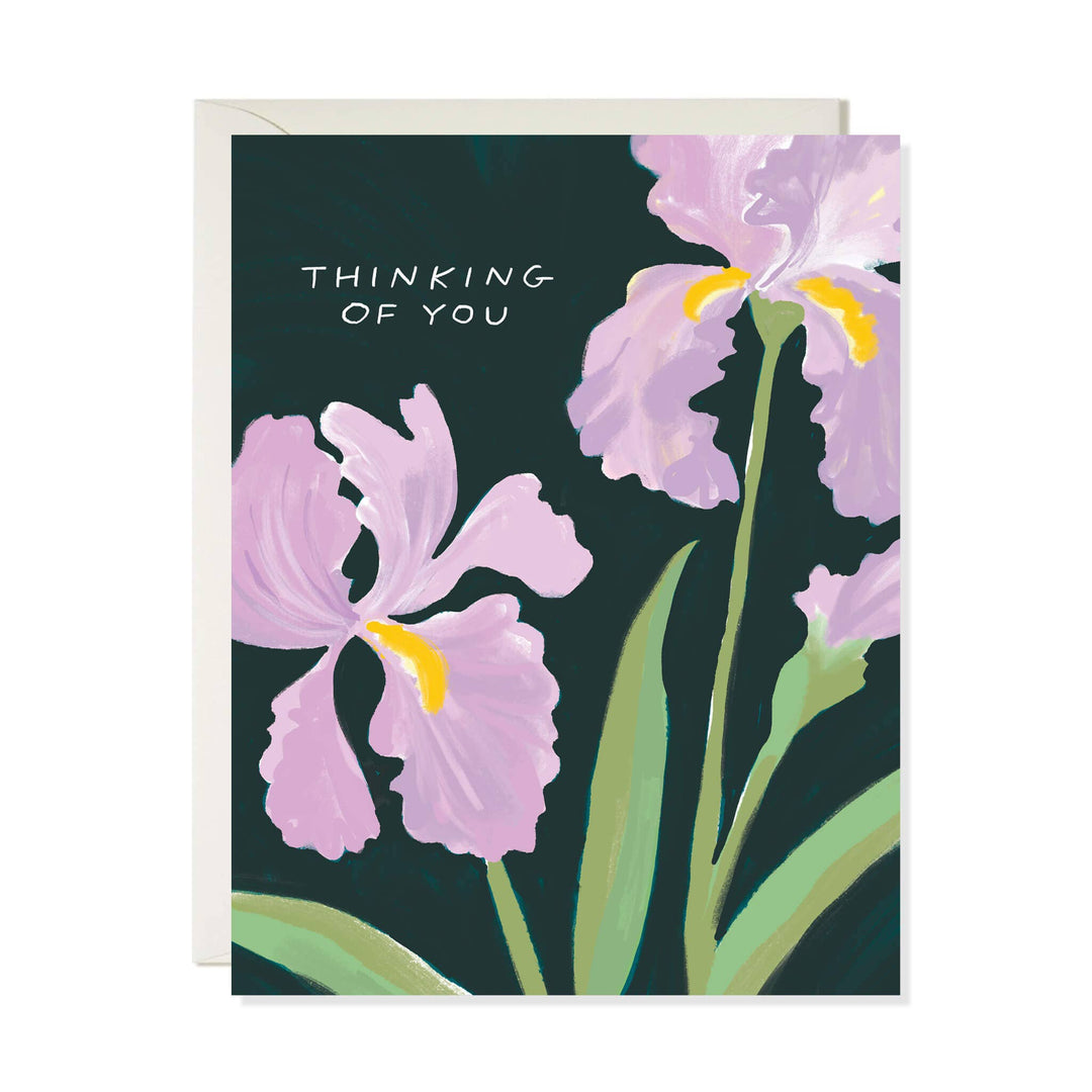 A greeting card featuring a dark green background with painted light purple irises and the words 'THINKING OF YOU' in white text, shown with a white envelope behind it.
