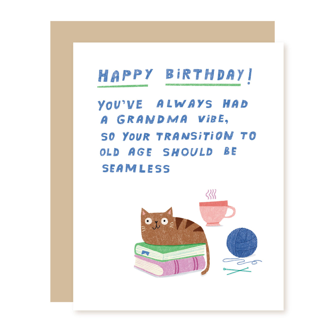 A birthday card with a tan envelope, featuring a white card with blue text that reads 'HAPPY BIRTHDAY! YOU'VE ALWAYS HAD A GRANDMA VIBE, SO YOUR TRANSITION TO OLD AGE SHOULD BE SEAMLESS.' Below the text is an illustration of a brown cat sitting on stacked books next to a pink cup, a blue ball of yarn, and knitting needles.