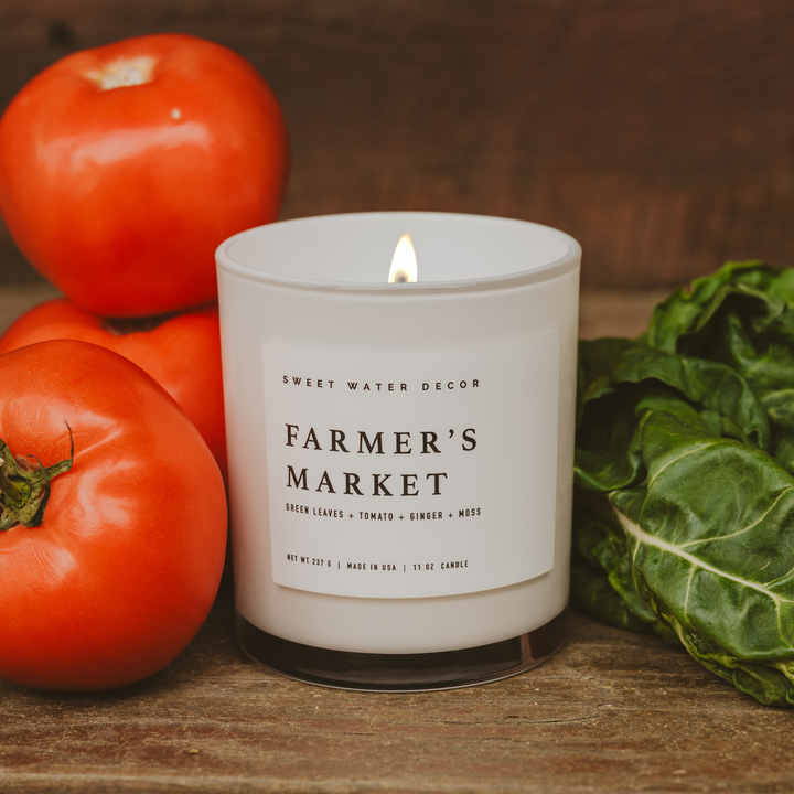 Candle in opaque white container with white label nestled between tomatoes and Swiss Chard leaves.