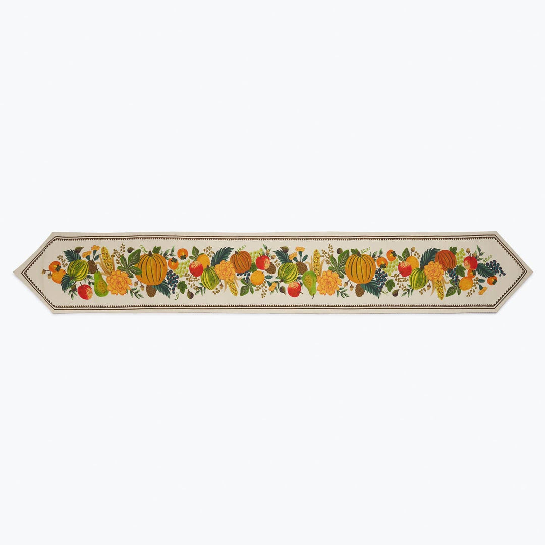 A long decorative table runner with an autumnal harvest design, featuring pumpkins, gourds, apples, and leaves on a cream background with a dark border.