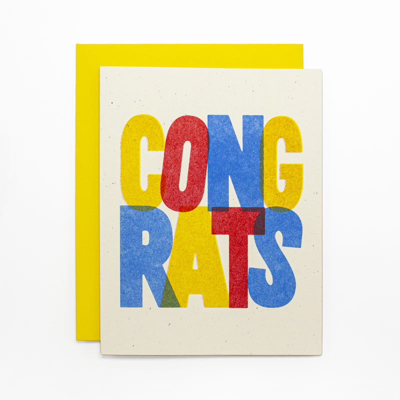 A greeting card with a bright yellow envelope behind it, featuring the word 'CONGRATS' in large, overlapping multicolor letters on a cream background.