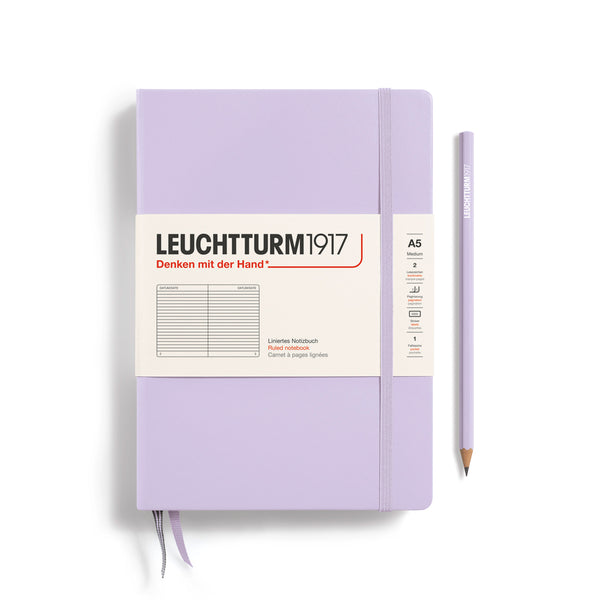 A Leuchtturm1917 A5 hardcover ruled page notebook in lilac with a white label on the front, shown closed with an elastic closure and ribbon bookmark. A matching lilac pencil is placed vertically to the right.