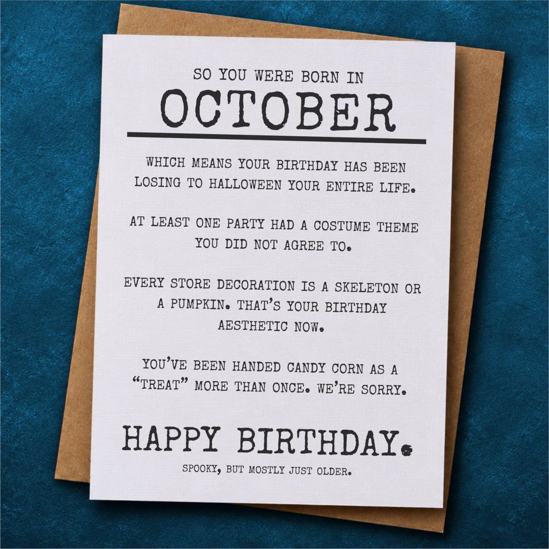 A white birthday card with black text that reads 'SO YOU WERE BORN IN OCTOBER' and a spooky message about Halloween costume themes, placed on top of a brown kraft envelope.