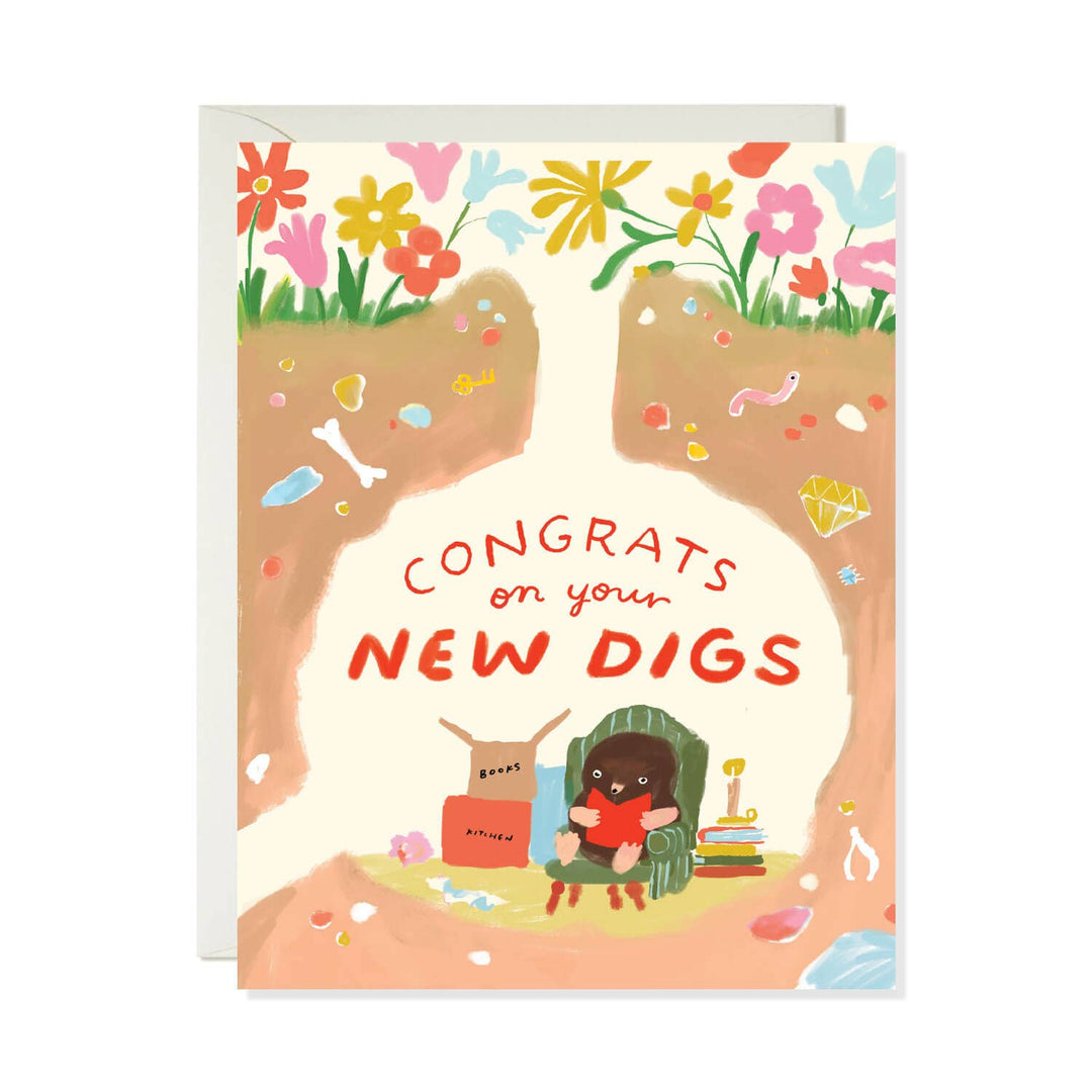 A greeting card with a whimsical illustration of a mole sitting in a green chair inside a cave, with the text 'CONGRATS on your NEW DIGS' in red. The card is shown with a white envelope behind it.