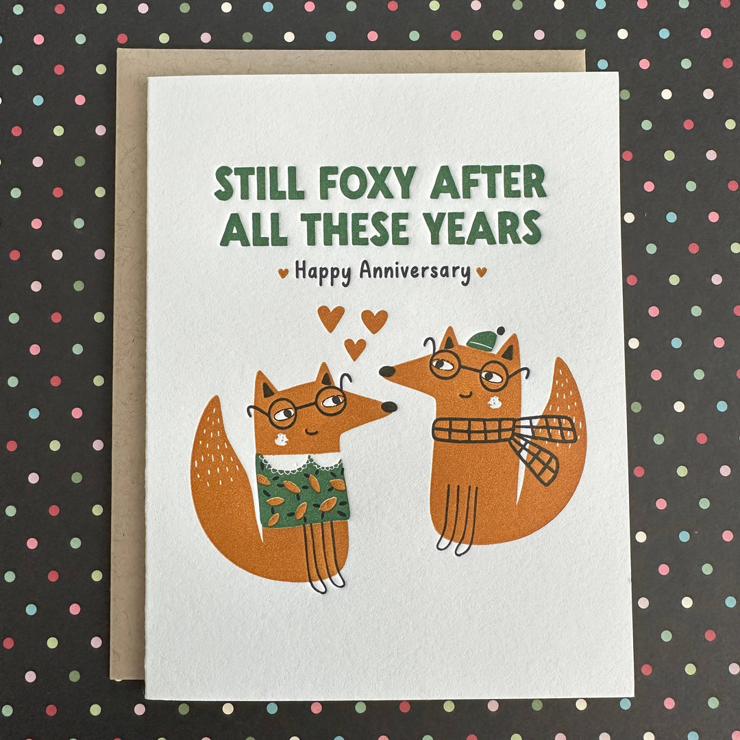 Two hand drawn foxes wearing glasses and winter wear, with three small hearts between them. Green text on white card stock