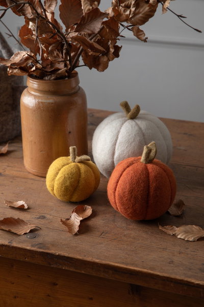 Felt pumpkins in orange, yellow, and white on a wooden surface with a vase of dried leaves.