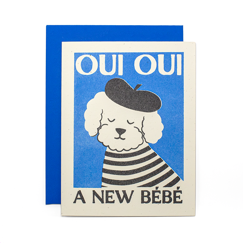 A greeting card with a blue envelope behind it, featuring a stylized dog illustration wearing a beret and striped shirt on a blue background, with the text 'OUI OUI' at the top and 'A NEW BÉBÉ' at the bottom.