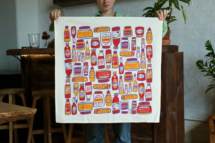 All the expected condiments drawn in reds and yellows on a white cotton kitchen towel.