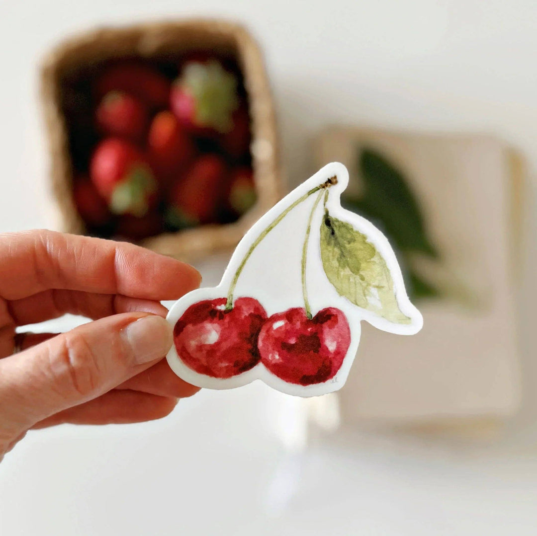 Hand holding sticker of two red cherries on stems with one leaf. A basket of cherries is in the background.