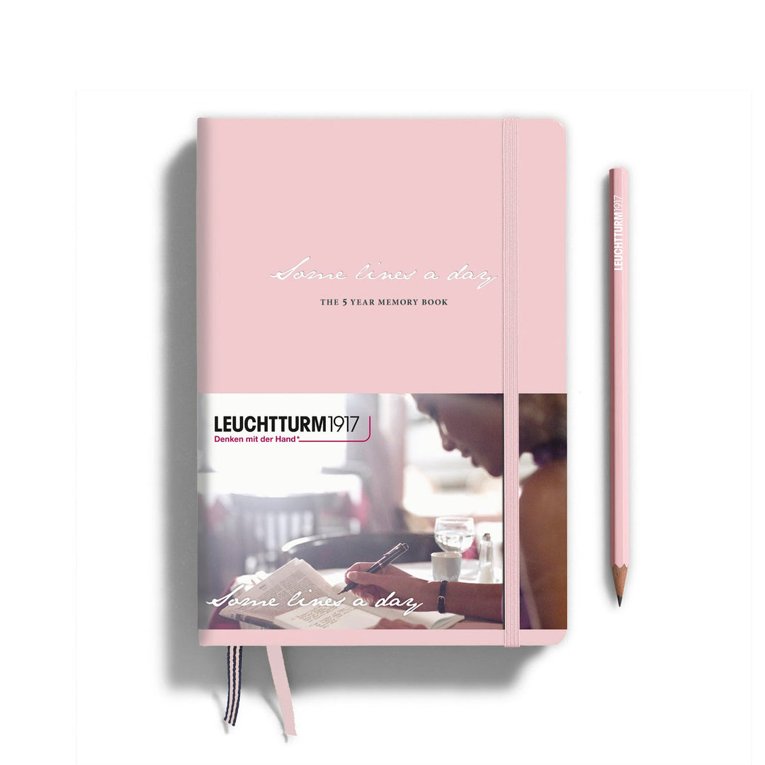 A pink hardcover memory book from Leuchtturm1917 with an elastic closure and a printed cover wrapper, shown alongside a matching pink Leuchtturm1917 pencil.