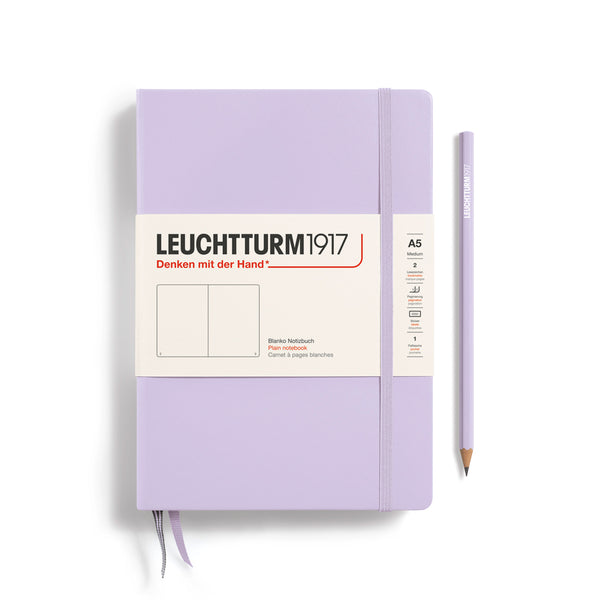 A Leuchtturm1917 A5 hardcover plain page notebook in lilac with a white label on the front, shown closed with an elastic closure and ribbon bookmark. A matching lilac pencil is placed vertically to the right.