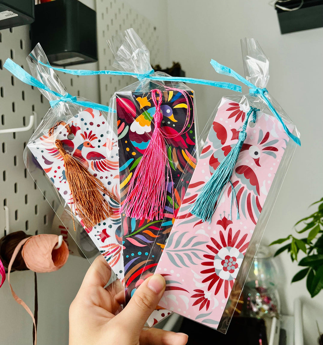 Hand holding three colorful bookmarks: white background with bronze tassel; black background with pink tassel; and pink background with blue tassel.