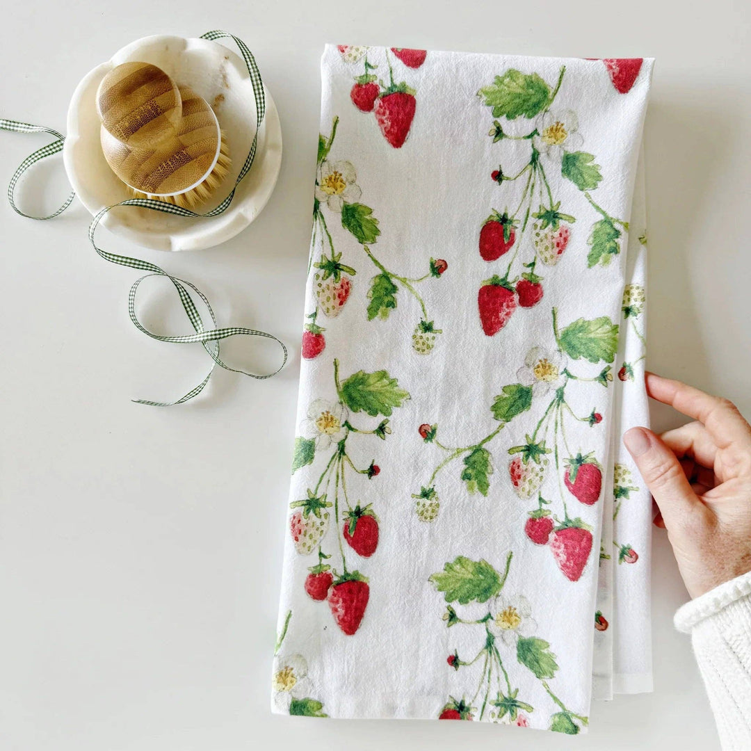 Hand holding a kitchen towel with a pattern of stems with ripe and unripe strawberries next to a kitchen bowl.