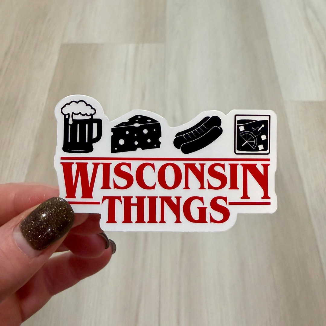 A hand with glittery brown nail polish holds a die-cut sticker that reads 'WISCONSIN THINGS' in red letters under black icon illustrations of a beer mug, wedge of cheese, hot dog, and brandy old fashioned.