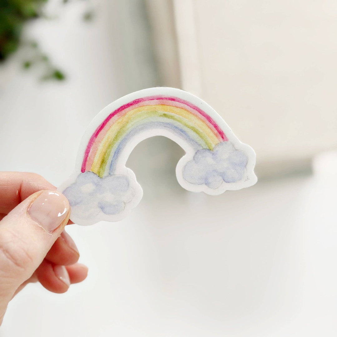 Hand holding a rainbow-shaped sticker with a cloud at each end, on a blurred background.