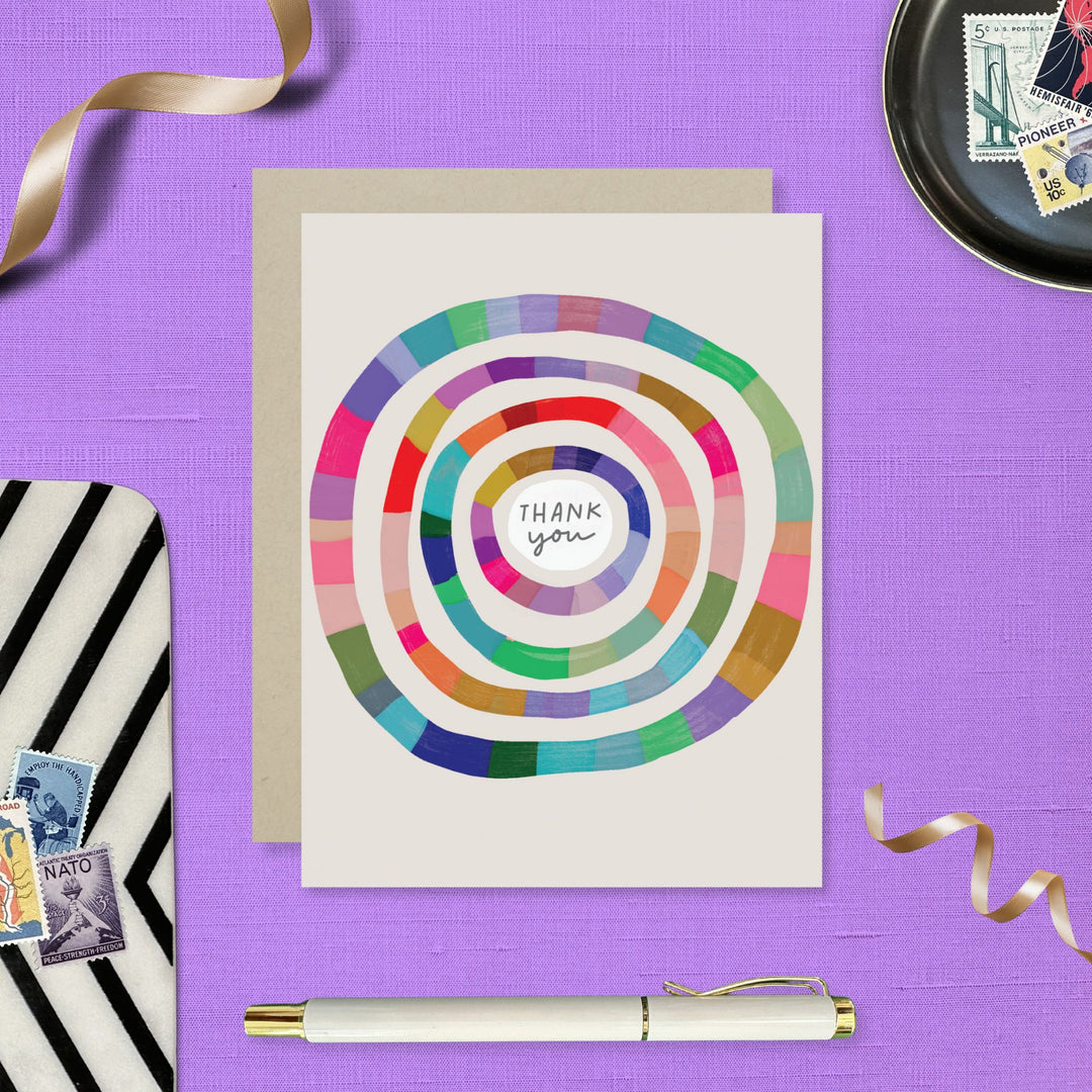 Front of card with multi-colored concentric circles on an off-white background, and the text "thank you" in the centermost circle. A pen and other table-top items are in view.