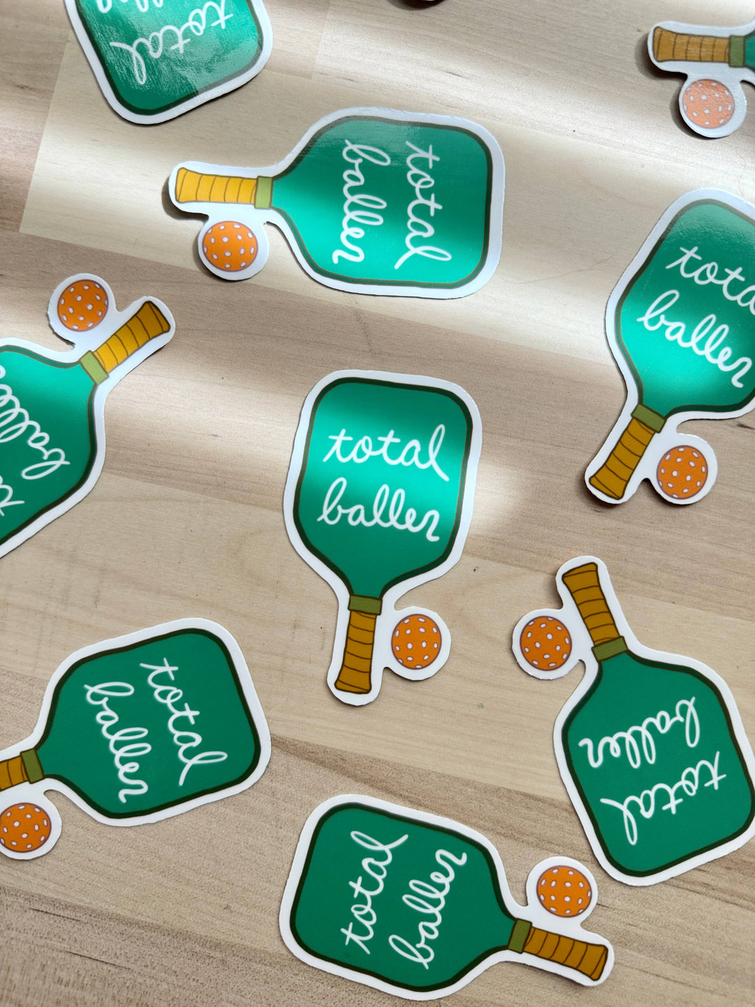 Several stickers on a table. Each sticker is a green pickleball racquet with "total baller" written in white. An orange ball is next to the grip.