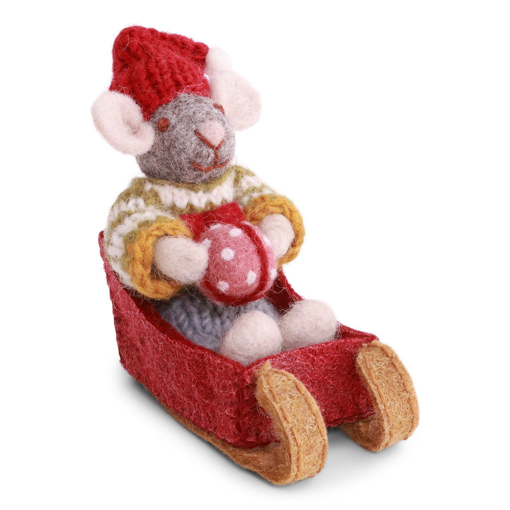 A handmade felt mouse wearing a red knit hat and a cream, yellow, and brown knit sweater, sitting in a red felt sleigh and holding a red ornament with white polka dots/