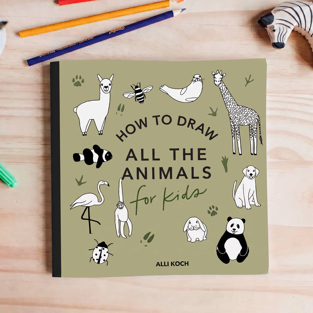 The book 'How to Draw All the Animals for Kids' by Alli Koch, featuring a sage green cover with black and white animal illustrations, displayed on a light wood desk with drawing pencils and a zebra toy.