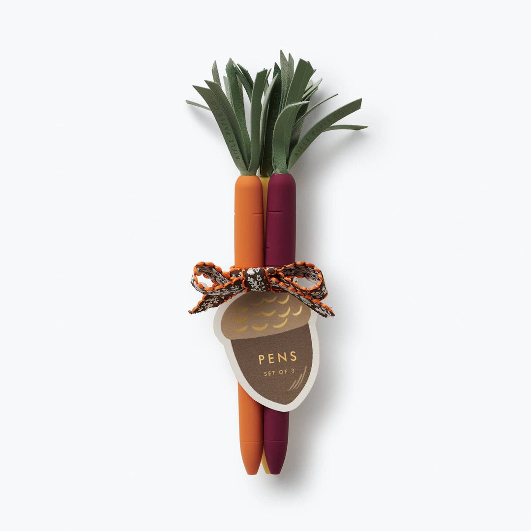 Carrot-shaped pen set in white, orange and red, with green tassels at the top and tied with a brown and orange fabric bow and an acorn-shaped label with the word 'pens.'