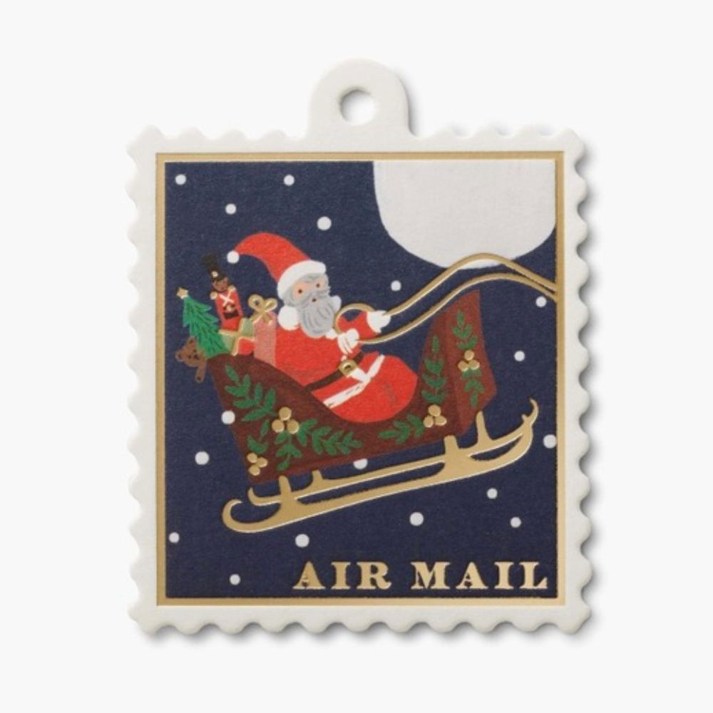 A postage stamp-style tag with a scalloped white border, featuring Santa Claus in a sleigh under a full moon with the words “AIR MAIL” at the bottom.
