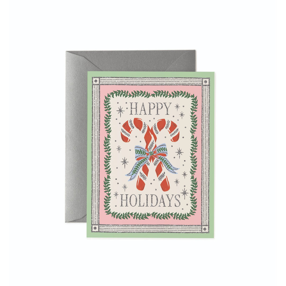A  Christmas greeting card with a gray envelope behind it, featuring the words “HAPPY HOLIDAYS” around an illustration of two candy canes tied with a blue bow, bordered by green wreath branches.