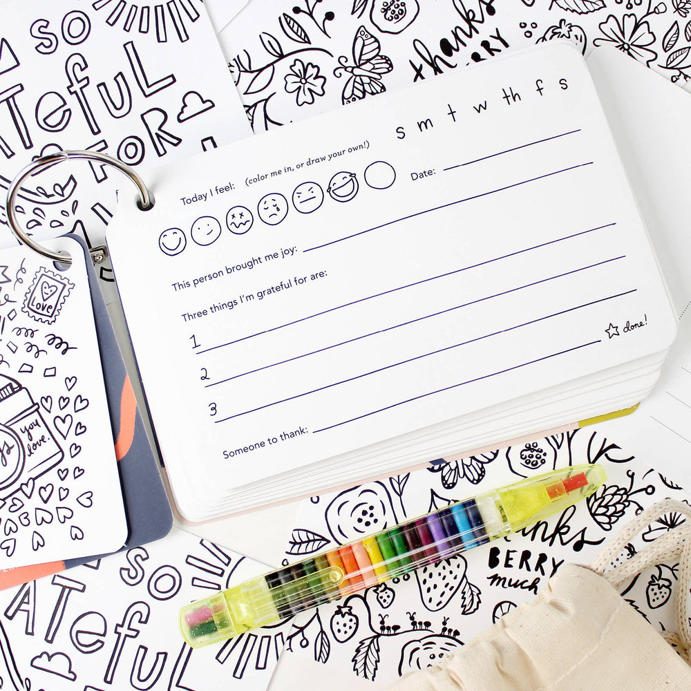 A page from the Kids Gratitude Kit, showin one of the pages in the journal, several of the color-in postcards, and the crayon pen.