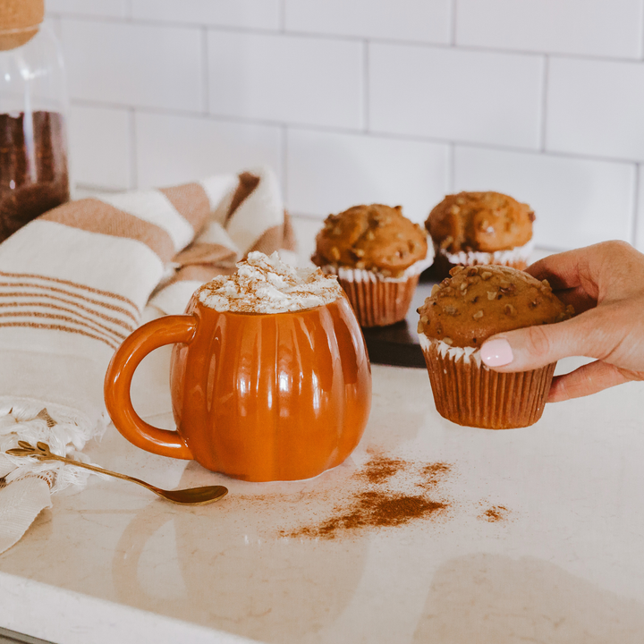 Orange mug in the shape of a pumpkin, topped with whipped cream, next to a person's hand holding a muffin.