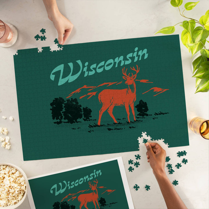 Hands of two people finishing the puzzle with 'Wisconsin' across the top and a drawing of a deer in a forest on a solid green background.