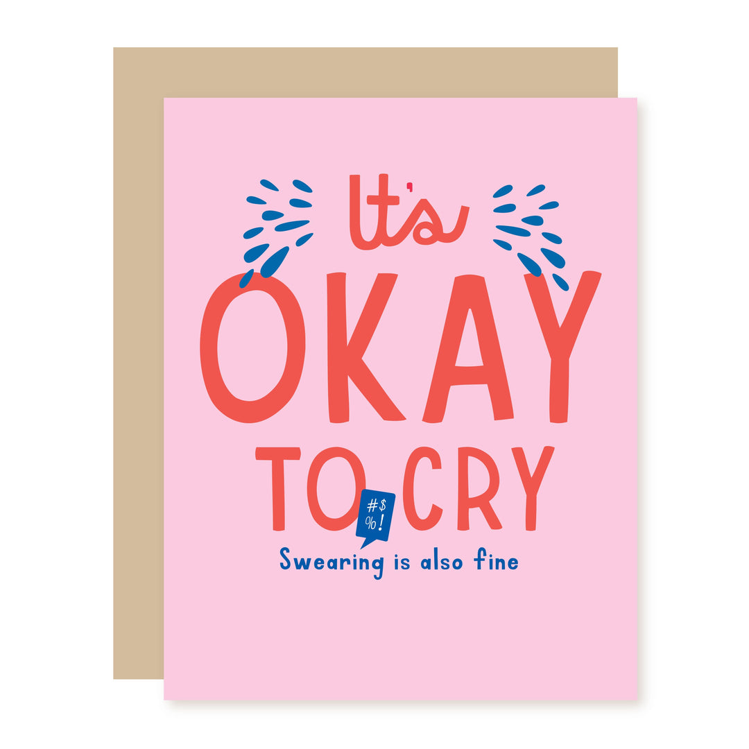 A pink greeting card with a beige envelope behind it. The card features the text 'It's OKAY TO CRY' in coral and blue lettering, with 'Swearing is also fine' written below in blue.