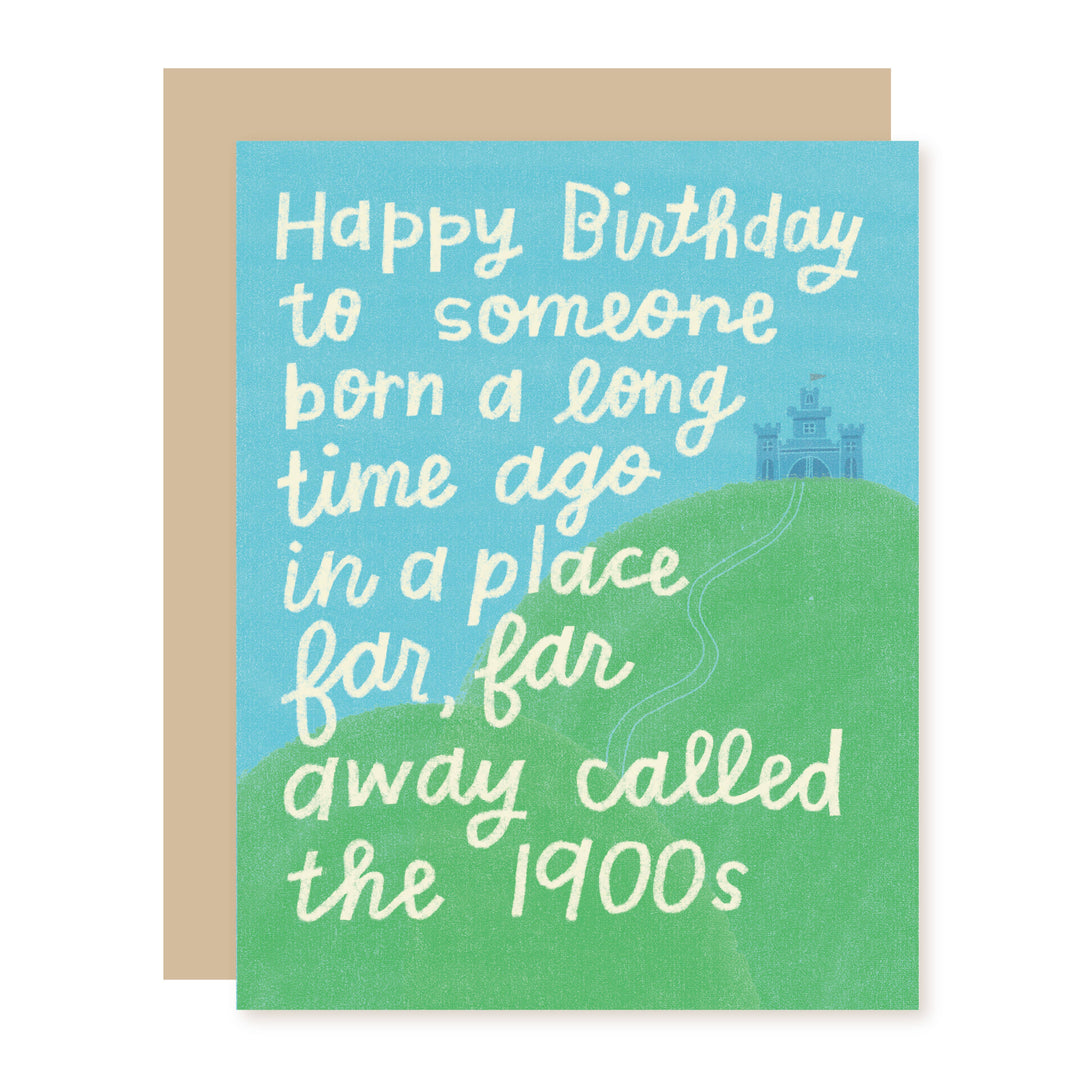 A square birthday card with a tan envelope behind it. The card has a light blue top section and a green bottom section with a small castle illustration, and white handwritten text that reads, 'Happy Birthday to someone born a long time ago in a place far, far away called the 1900s.