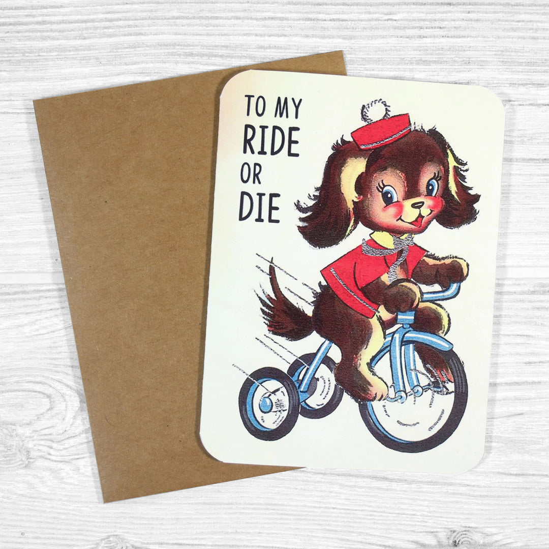 Greeting card with a dog riding a bike and 'To my ride or die' text, next to a brown envelope.