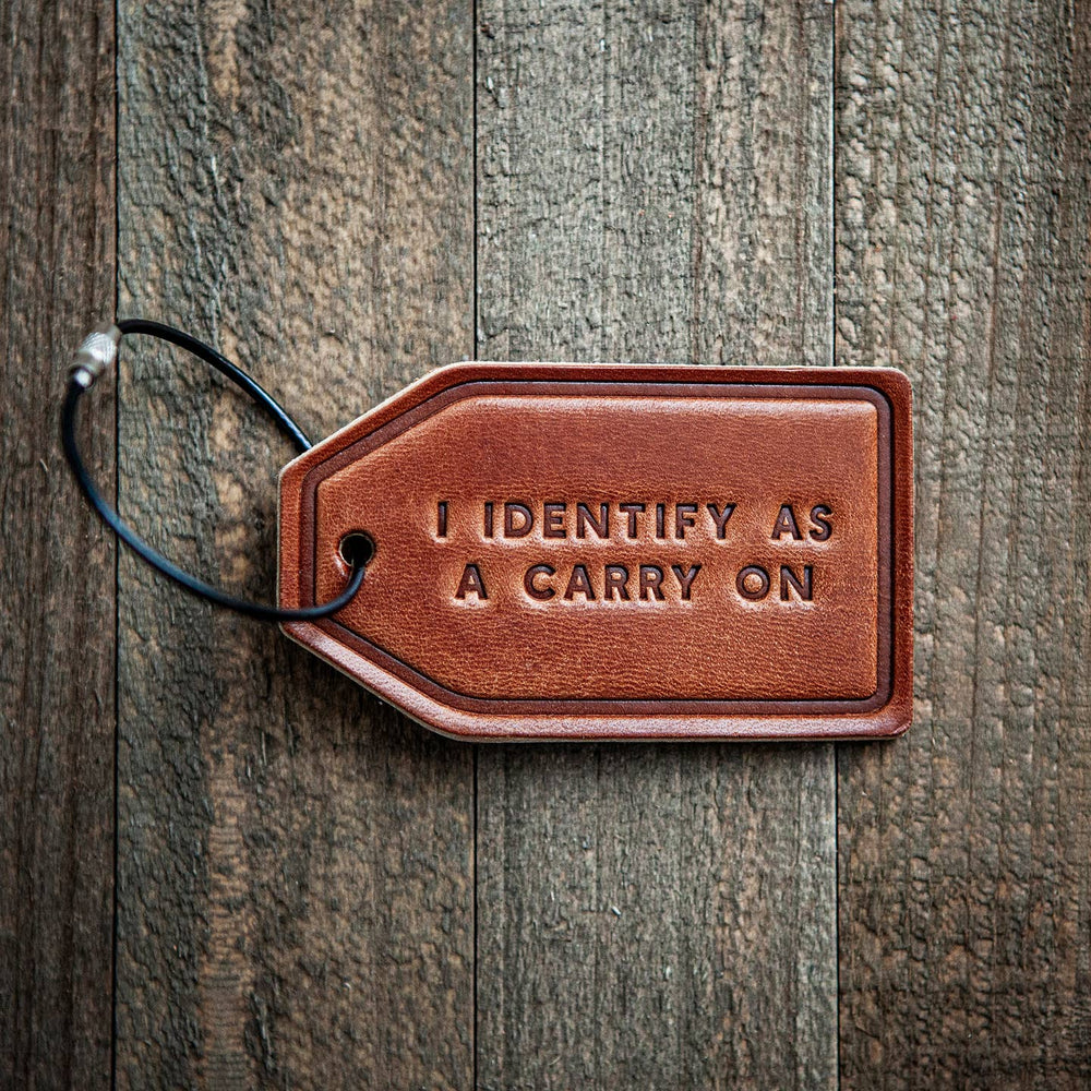 Brown leather luggage tag with 'I IDENTIFY AS A CARRY ON' text on a wooden surface
