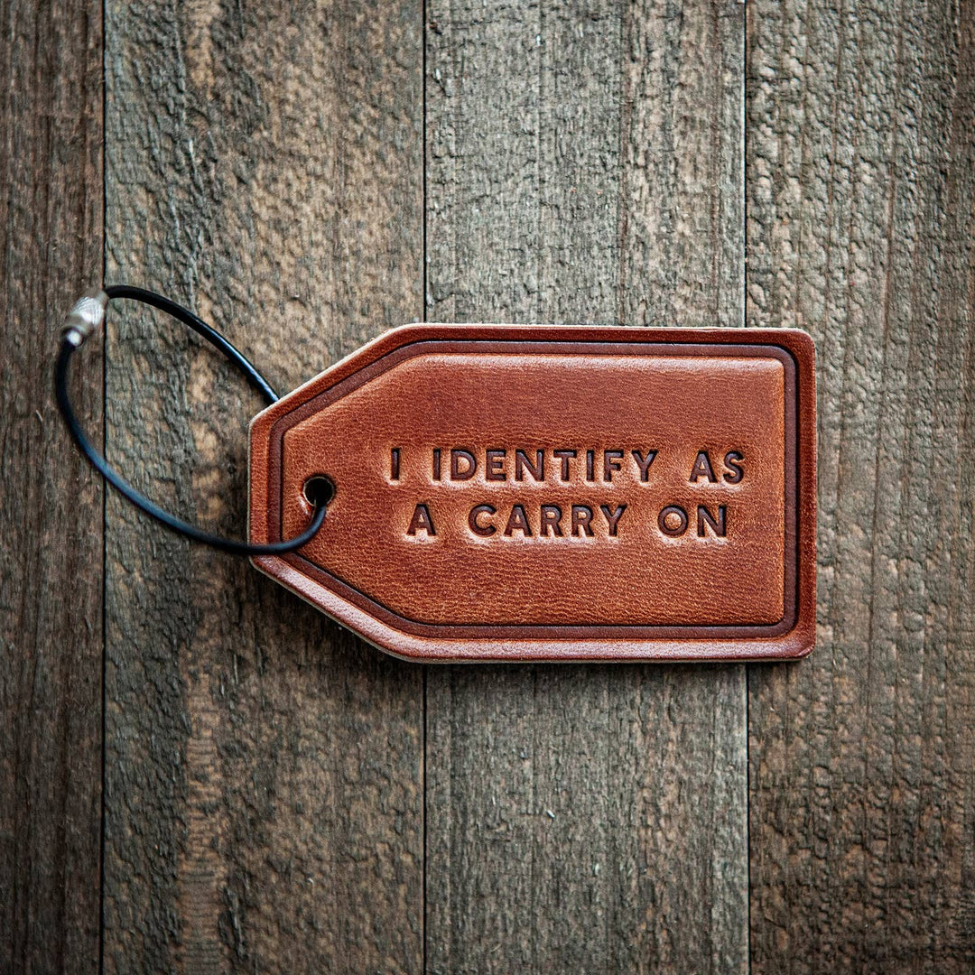 Brown leather luggage tag with 'I IDENTIFY AS A CARRY ON' text on a wooden surface