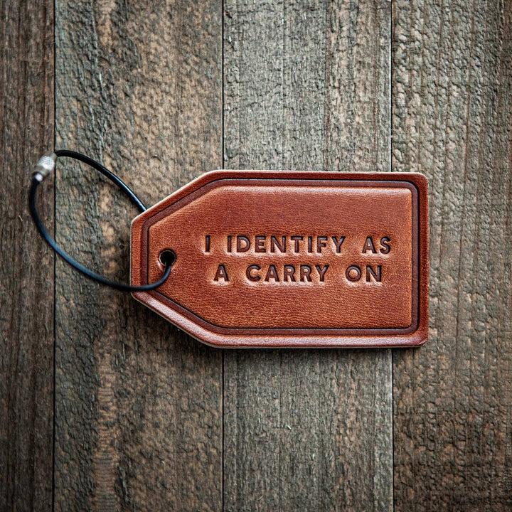 Brown leather luggage tag with 'I IDENTIFY AS A CARRY ON' text on a wooden surface
