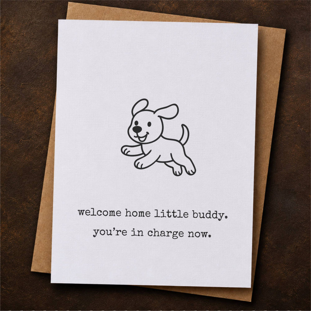 A white greeting card with a simple black line drawing of a puppy and the text 'welcome home little buddy, you're in charge now.' placed on top of a brown kraft envelope.