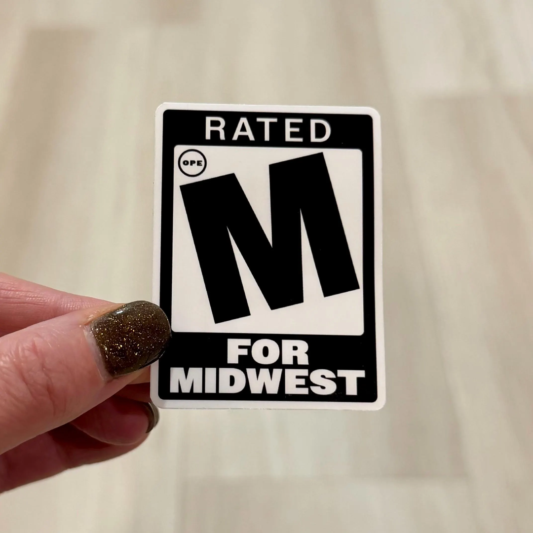 A hand with glittery brown nail polish holds a rectangular sticker featuring a large M and small 'ope' in a circle, with the text 'Rated M for Midwest' design.'