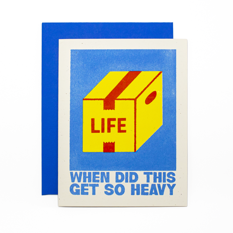 A greeting card with a white border and a blue backing card, featuring a yellow box labeled 'LIFE' with red tape on a blue background and the text 'WHEN DID THIS GET SO HEAVY' in blue at the bottom.