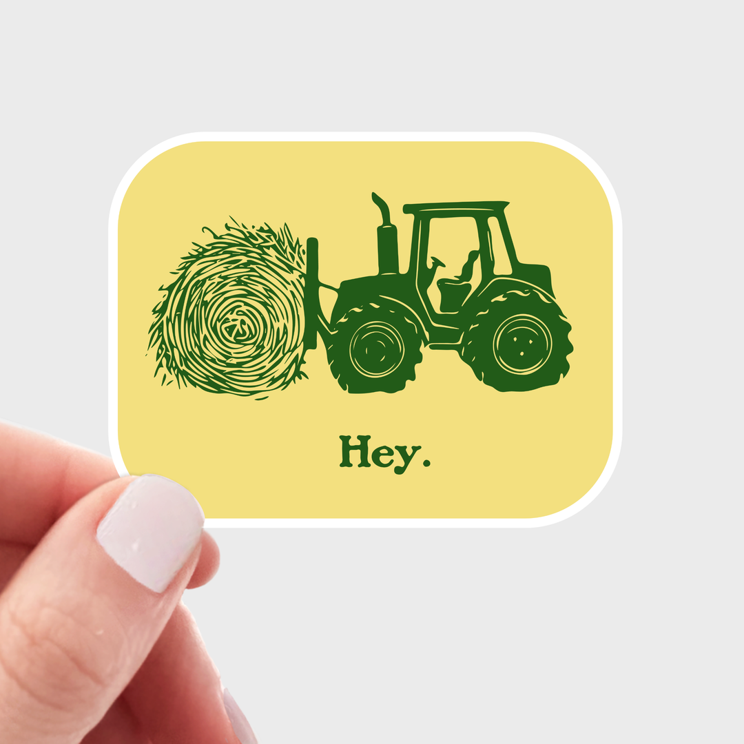 Hand holding a sticker with a green tractor and hay bale design on a yellow background.