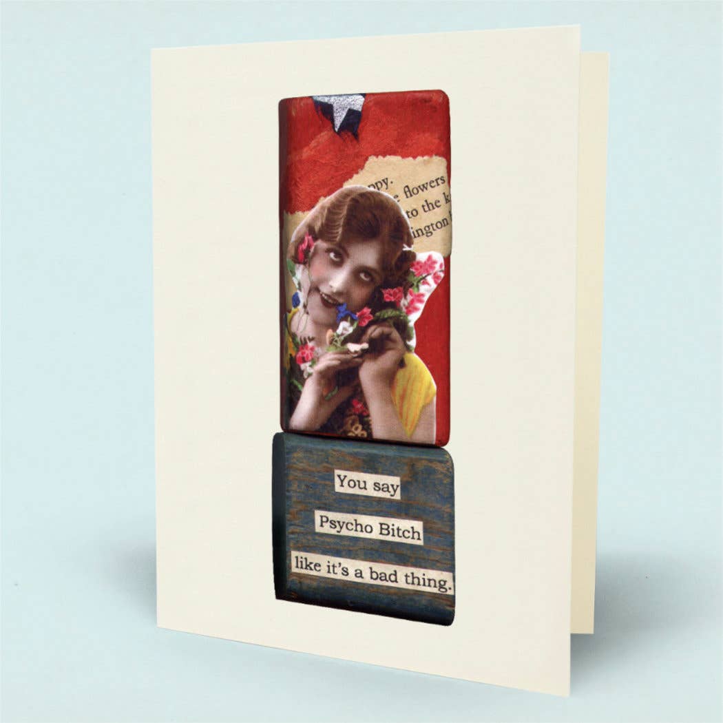 A greeting card shown standing upright. The card has an off-white border and features a collage-style front design with a vintage photo of a young girl holding flowers, a red textured panel, and a dark blue wood-textured section with the phrases 'You say Psycho Bitch like it's a bad thing'