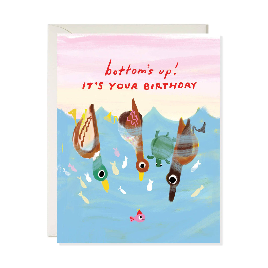 A greeting card with a whimsical illustration of three ducks upside down in blue water under a pink sky, with the text 'bottom's up! IT'S YOUR BIRTHDAY' in red. A white envelope is partially visible behind the card.