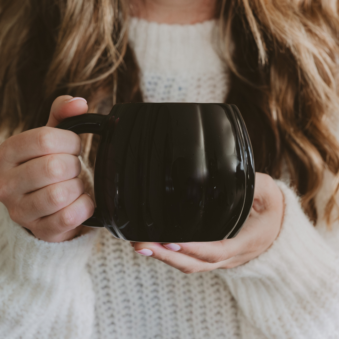 Person holding a black mug in the shape of a pumpkin, with a blurred background