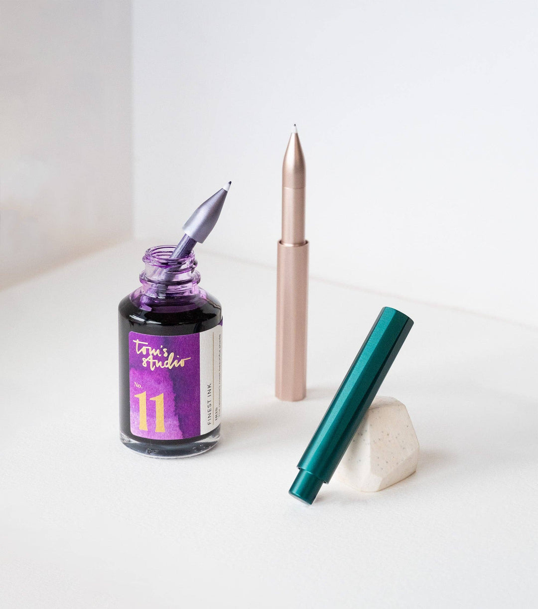 Pieces of the pen: a silver one in a bottle of ink, a blush one standing upright and the barrel of a green one leaning on a support.