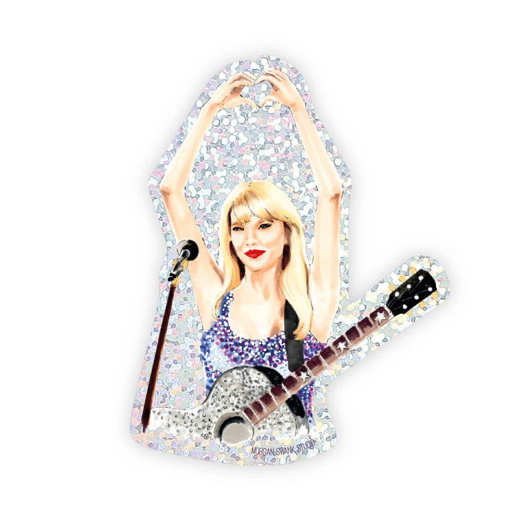 Taylor Swift at the microphone with a guitar, making a heart sign with her hands held above her head. and surrounded by glitter.