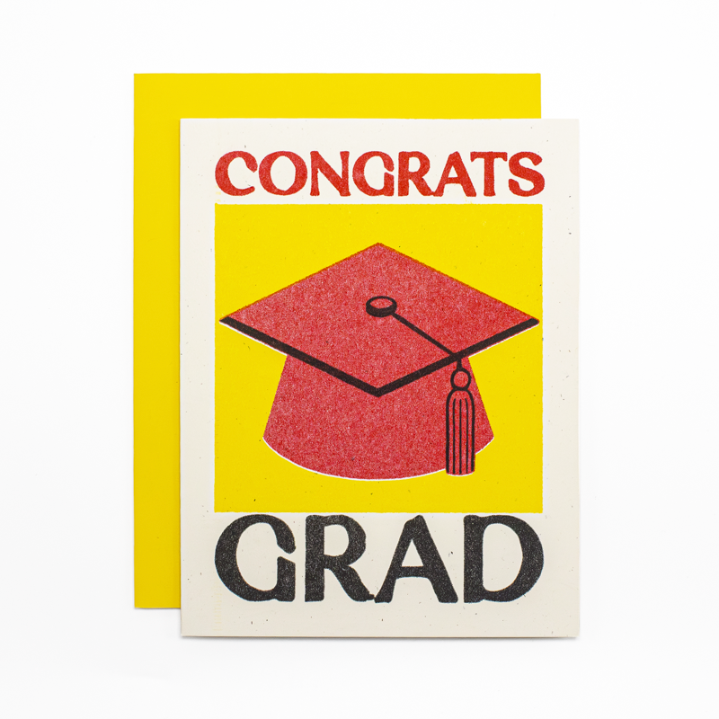 A graduation card with a bright yellow envelope behind it. The card has an off-white border and reads 'CONGRATS GRAD' in red and black text, with a red graduation cap illustration in the center.