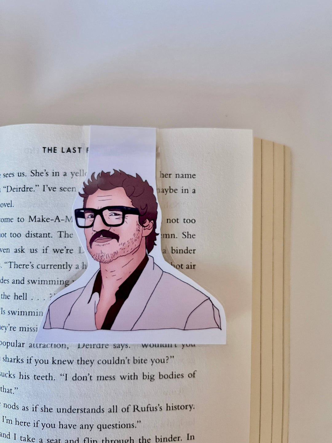Bookmark with drawing of Pedro Pascal wearing large black glasses and a black shirt with pink sportscoat shown clipped over a page in a book.