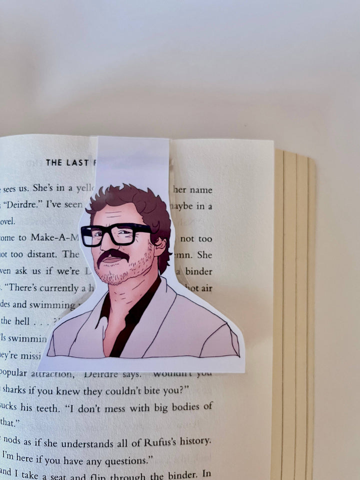 Bookmark with drawing of Pedro Pascal wearing large black glasses and a black shirt with pink sportscoat shown clipped over a page in a book.